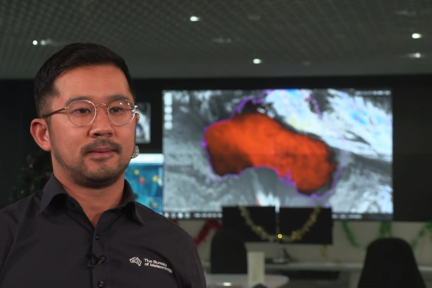A man with dark hair and glasses in a black collared shirt stands in front of a screen showing a graphic of Australia.