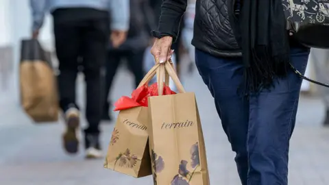 Bloomberg via Getty Images Cropped shot of the bottom half of a shopper wearing jeans and carrying two brown paper Terrain bags at Broadway Plaza in Walnut Creek, California, US, on Thursday, Dec. 11, 2025. 