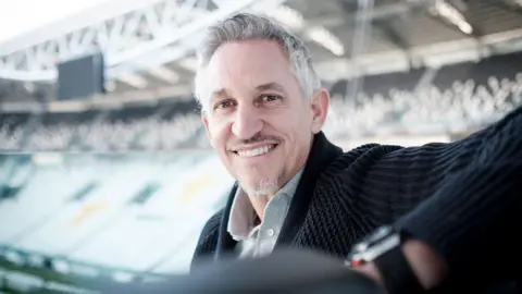 Juventus FC/Getty Images Gary Lineker sits in a football stand smiling, with another stand across the pitch seen behind him out of focus.