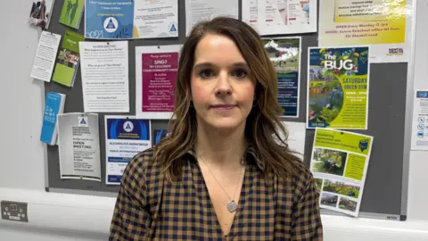 Joy Watson is wearing a brown and navy checked shirt. She has a silver chain around her neck and long brown hair. .She is standing in front of a notice board with lots of coloured posters on it.