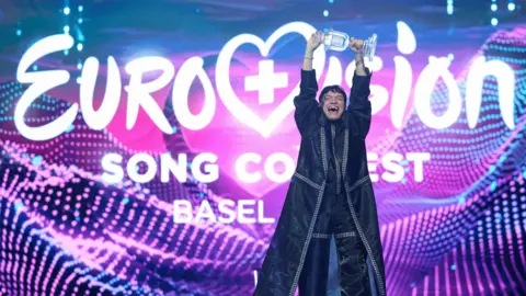 Reuters JJ holding aloft the trophy in celebration at Eurovision 2025 in front of a giant colourful Eurovision Song Contest branded backdrop
