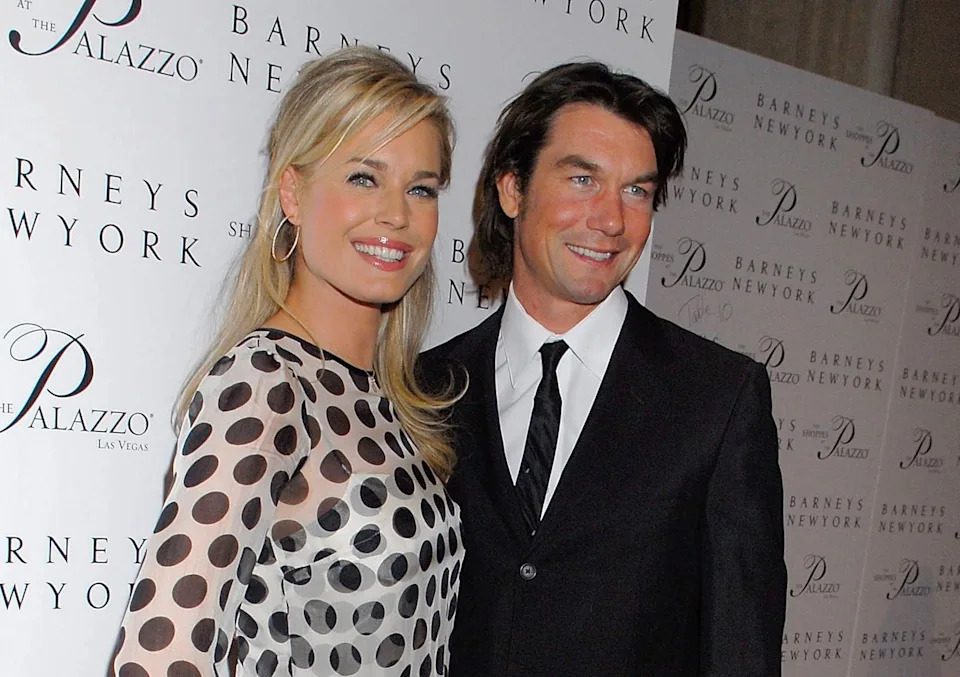 Two individuals pose on a red carpet. The woman wears a polka dot dress; the man wears a dark suit and tie. Background features event branding