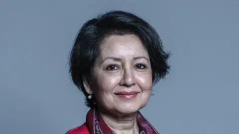 House of Lords Baroness Shas Sheehan, a woman with dark cropped hair and pearl earrings, smiles into the camera. 