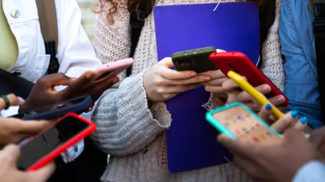 A circle of young people from the shoulders down holding a smartphones in multicoloured cases