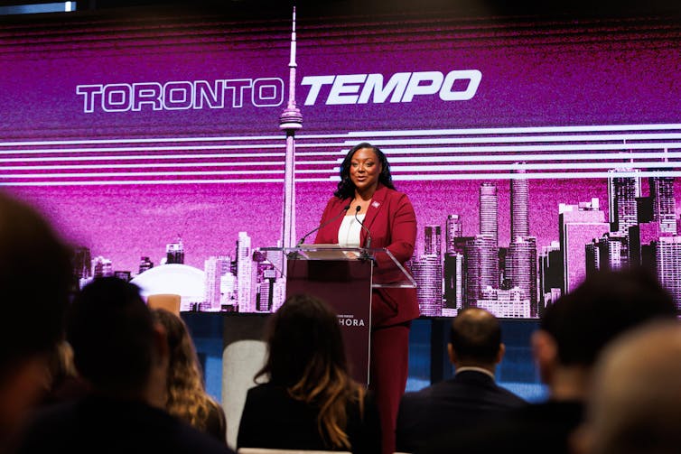 A Black woman in a red pantsuit speaks from behind a podium to a crowd of people. Behind her is an image of the Toronto skyline with the phrase 'Toronto Tempo' overlaid