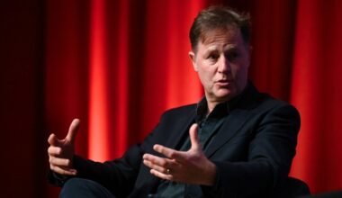 Nick Clegg gestures while speaking on stage against a red curtain backdrop.