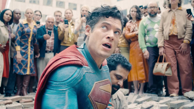 David Corenswet as Superman surrounded by a crowd as he's sitting in rubble in Superman