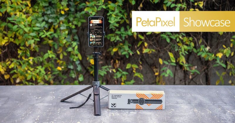 A smartphone mounted on a compact, extended tripod stands on a concrete surface outdoors. Next to it is the tripod's packaging box. In the background, leafy greenery and a "PetaPixel Showcase" banner are visible.