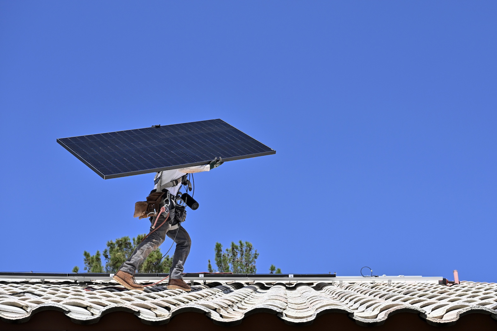 A person wearing jeans and a tool belt carries a solar panel across a roof, with a deep blue sky behind them