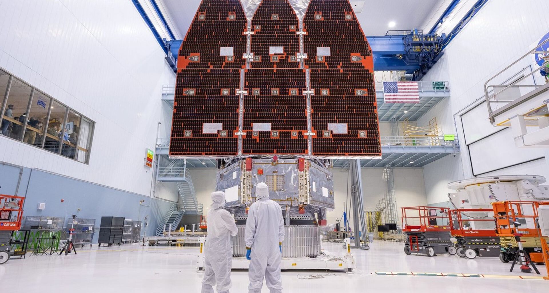 Three large solar panels hang in the back of a cleanroom warehouse room where two workers dressed in white suits stand in the foreground