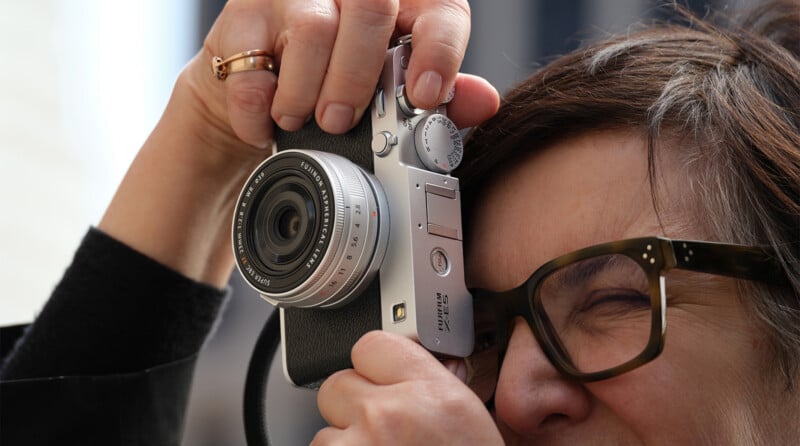 A person with short hair and glasses is holding a silver camera close to their face, looking through the viewfinder and preparing to take a photo.