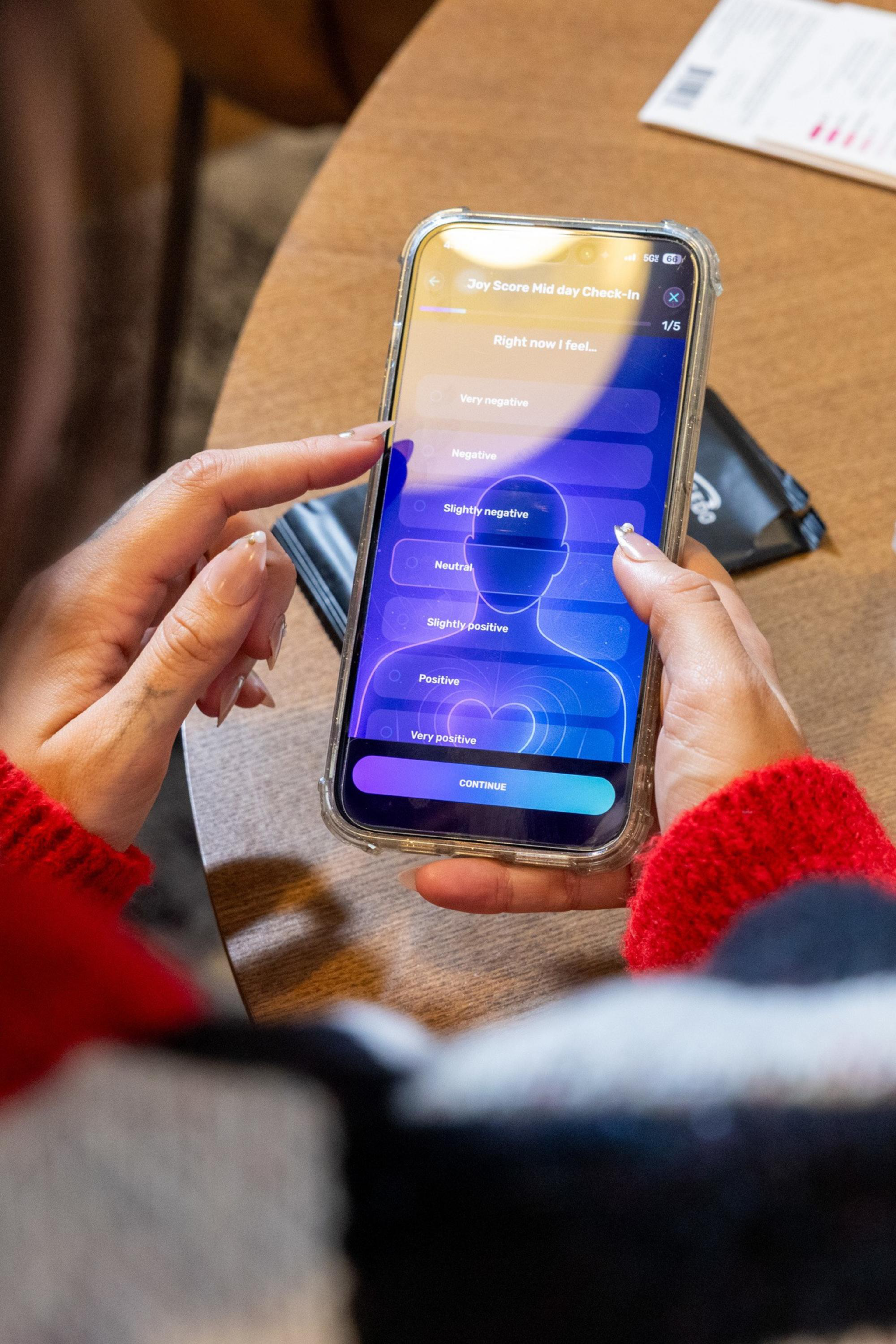 A person with long nails wearing a red sweater is using a smartphone app to select their current mood from options ranging from “Very negative” to “Very positive.”