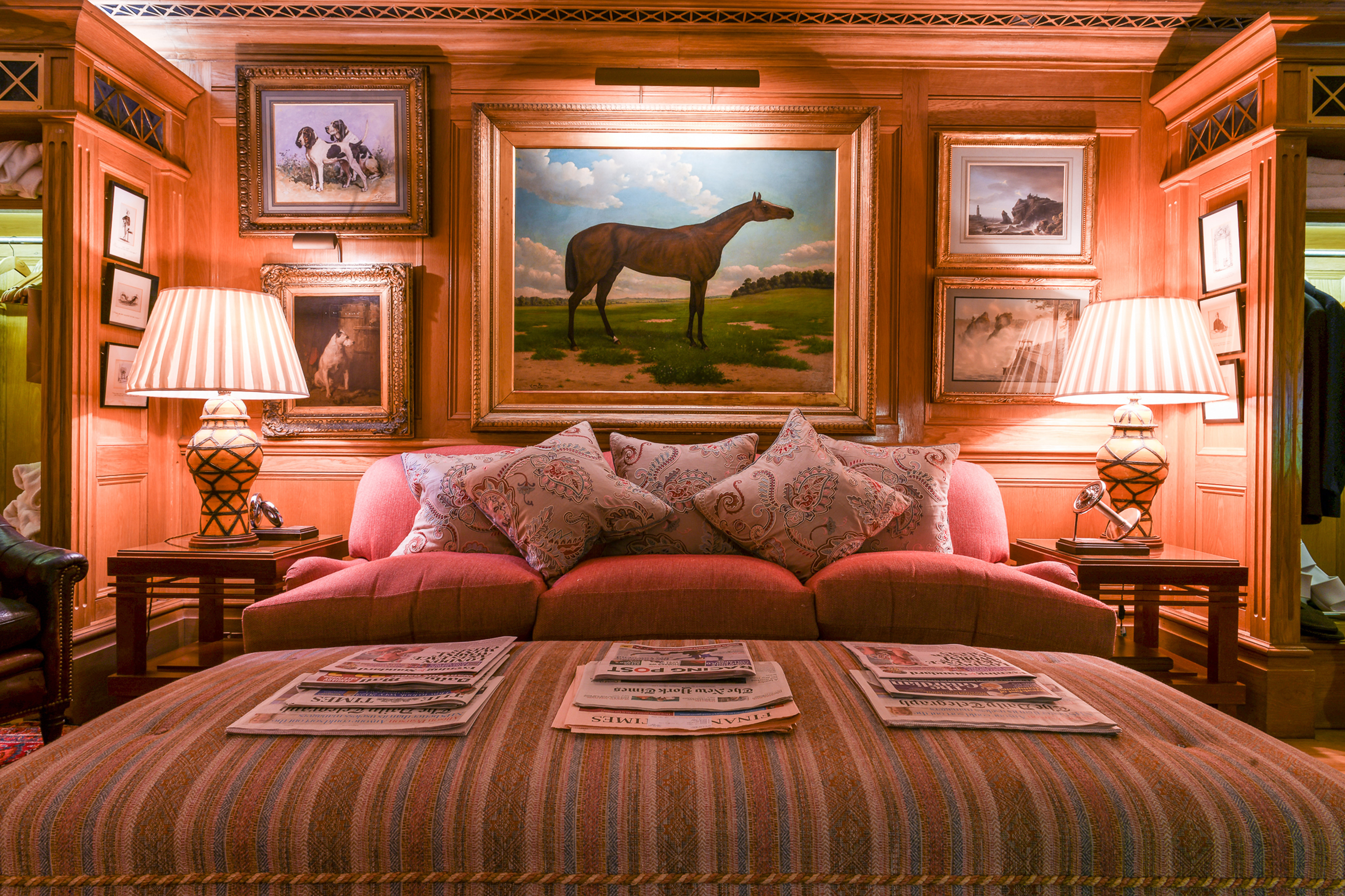 A cozy room with paintings of horses and dogs, a red sofa, and newspapers on an ottoman.