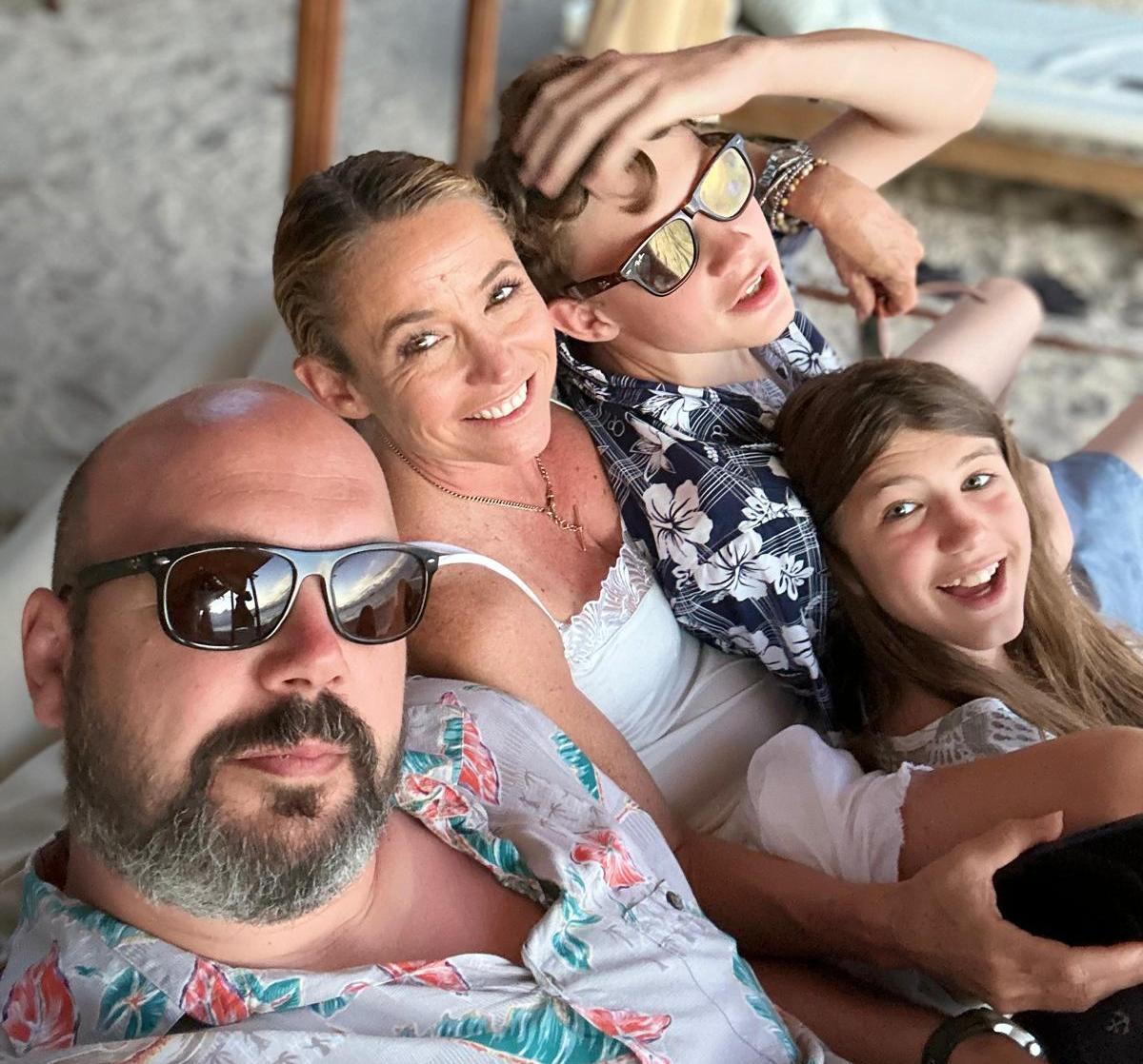 A family of four, including two children, wearing sunglasses and smiling while sitting on a beach.