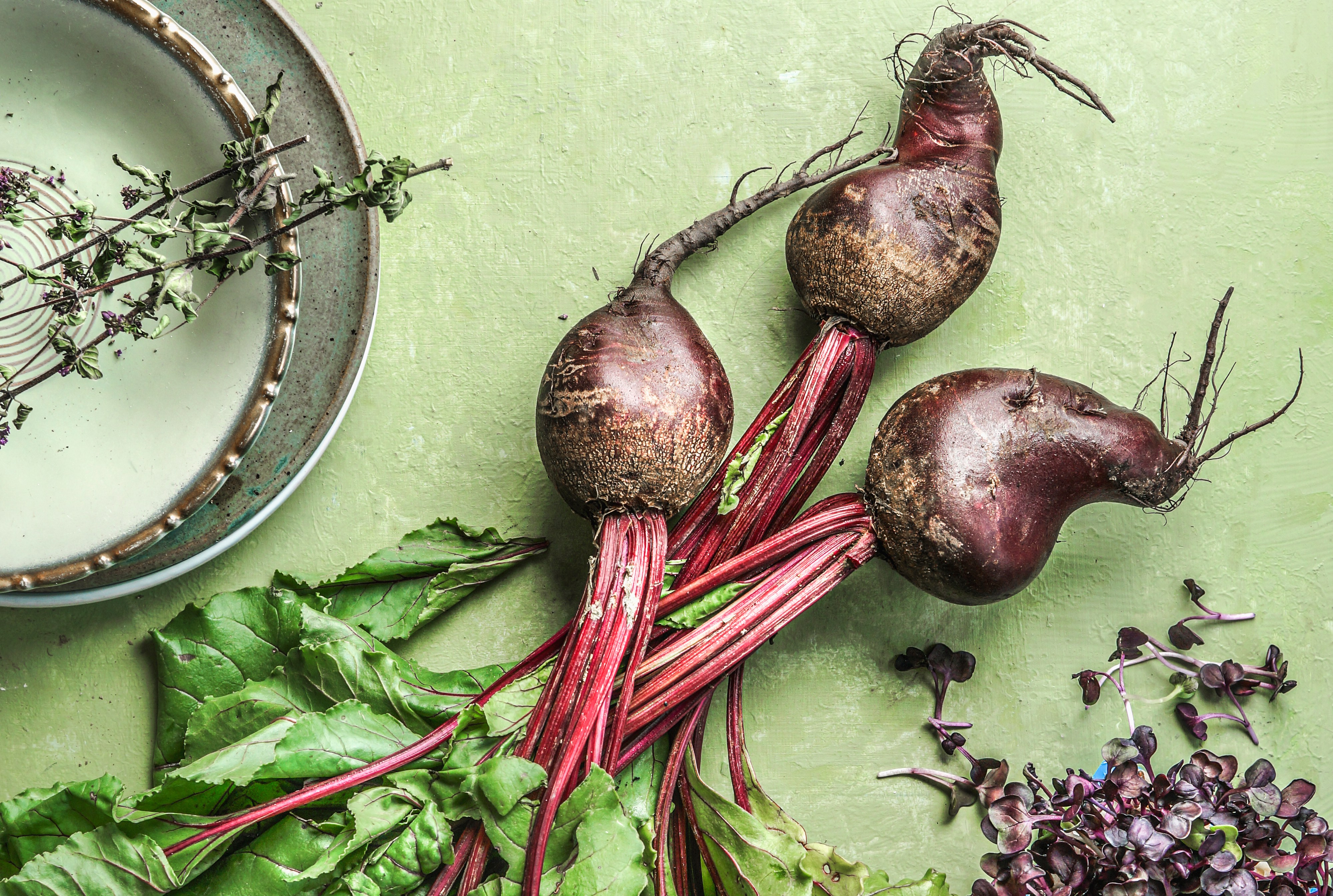 Top-down view of a bunch of raw organic beetroots with greens, next to ingredients like bay leaves and peppercorns, on a light green surface.