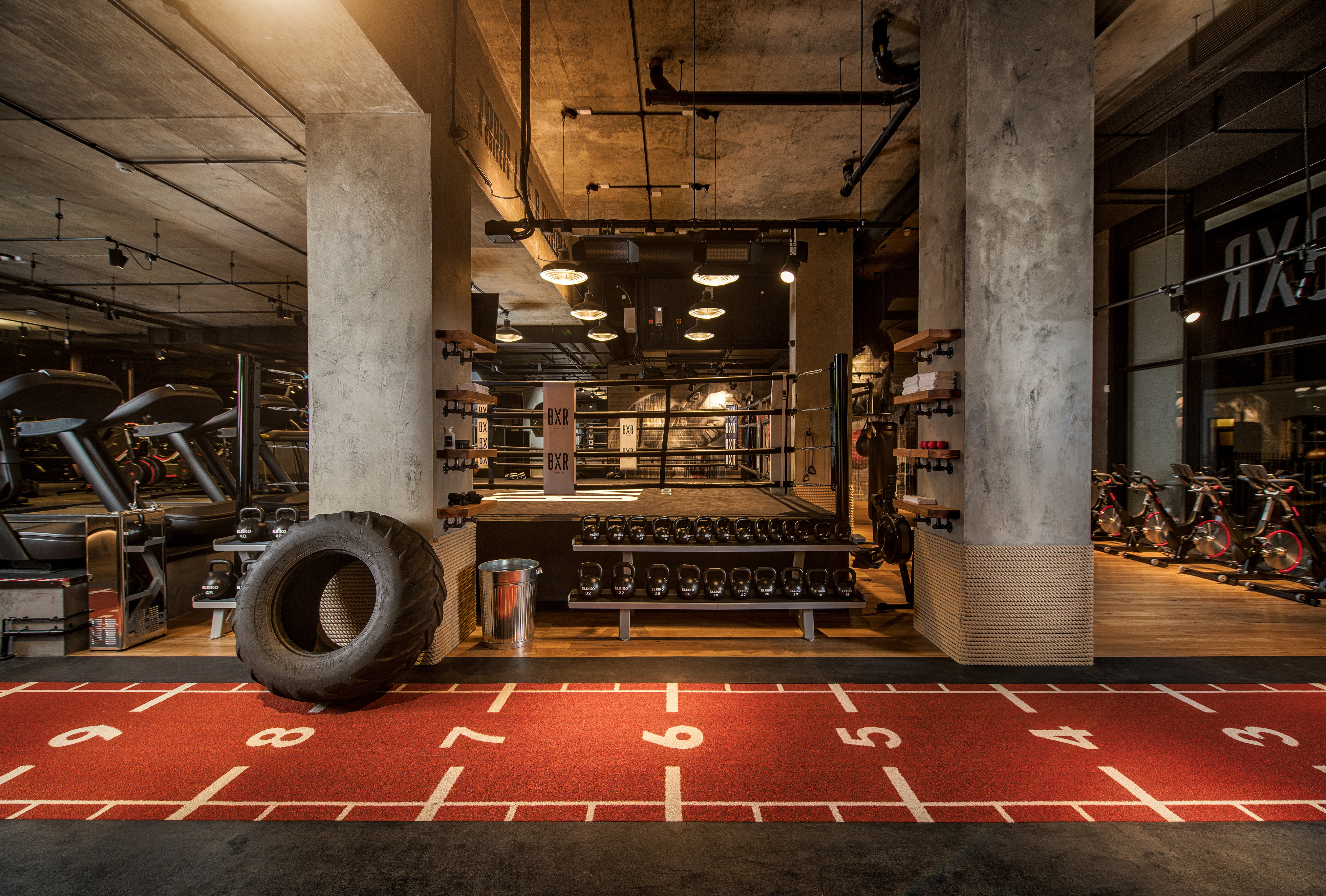 A boxing gym with a red running track in the foreground, a boxing ring in the center, and spinning bikes in the background.