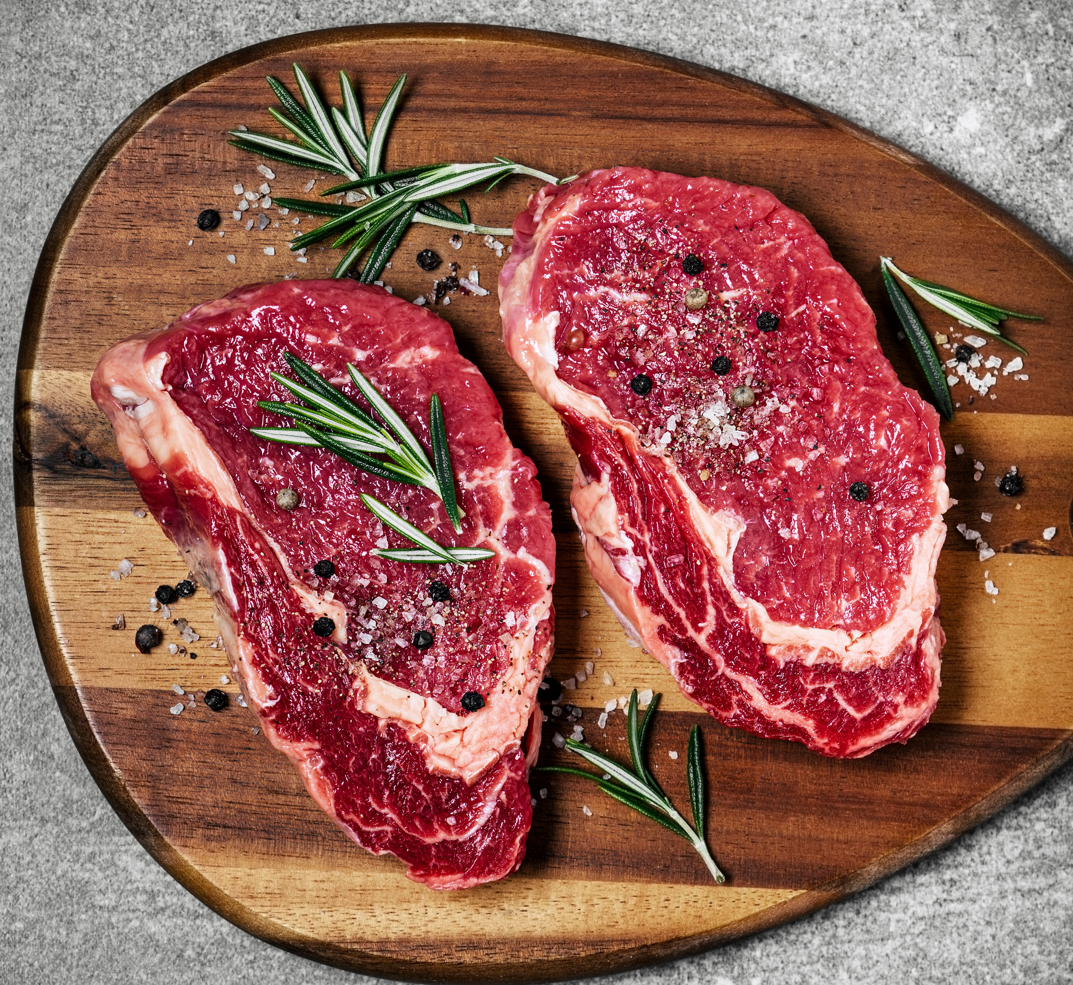 Two raw ribeye steaks on a wooden cutting board with rosemary, salt, and pepper.