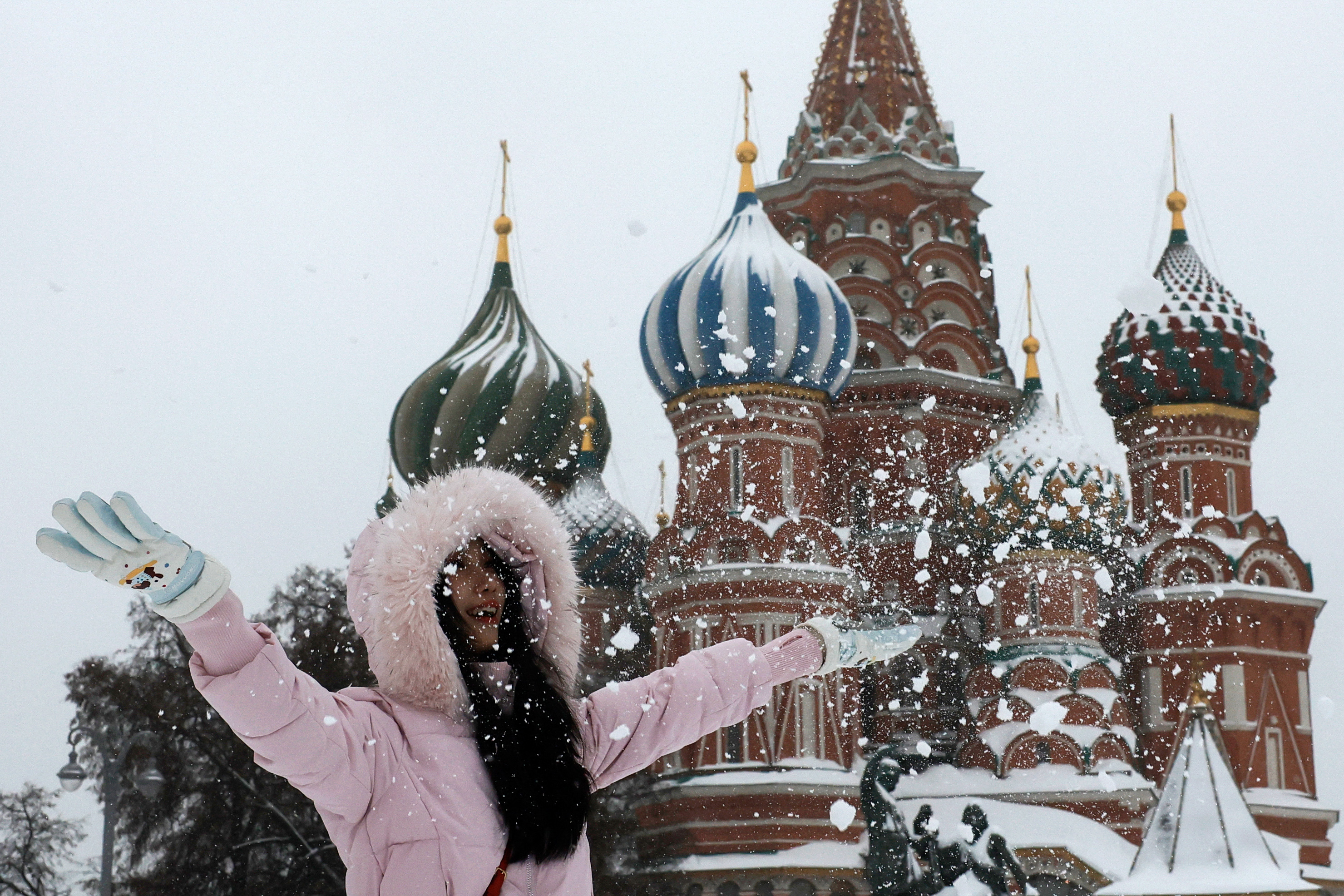 A woman in a pink coat with a fur-lined hood plays with snow in front of St. Basil's Cathedral.