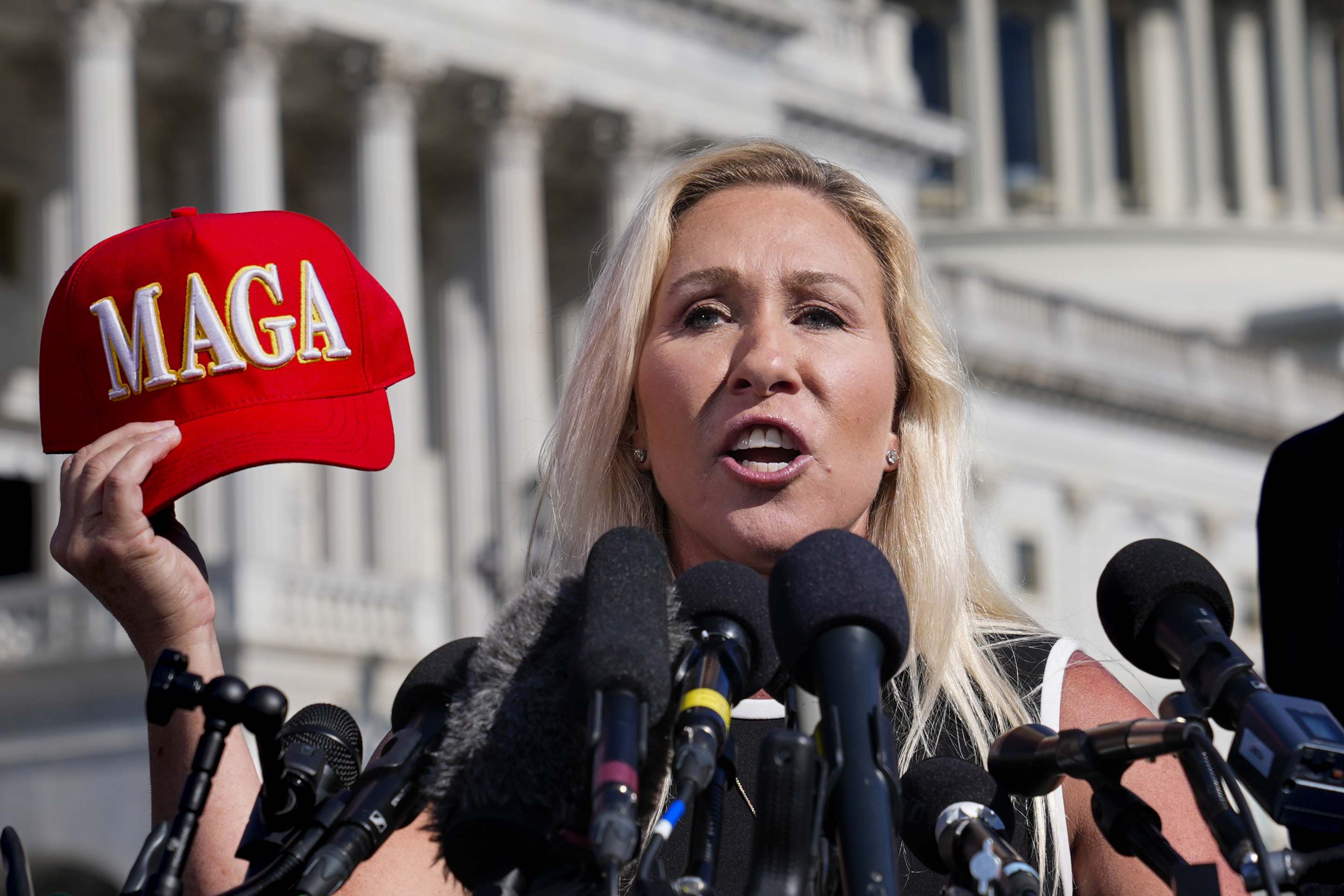 Representative Marjorie Taylor Greene holding a red "MAGA" hat at a news conference.