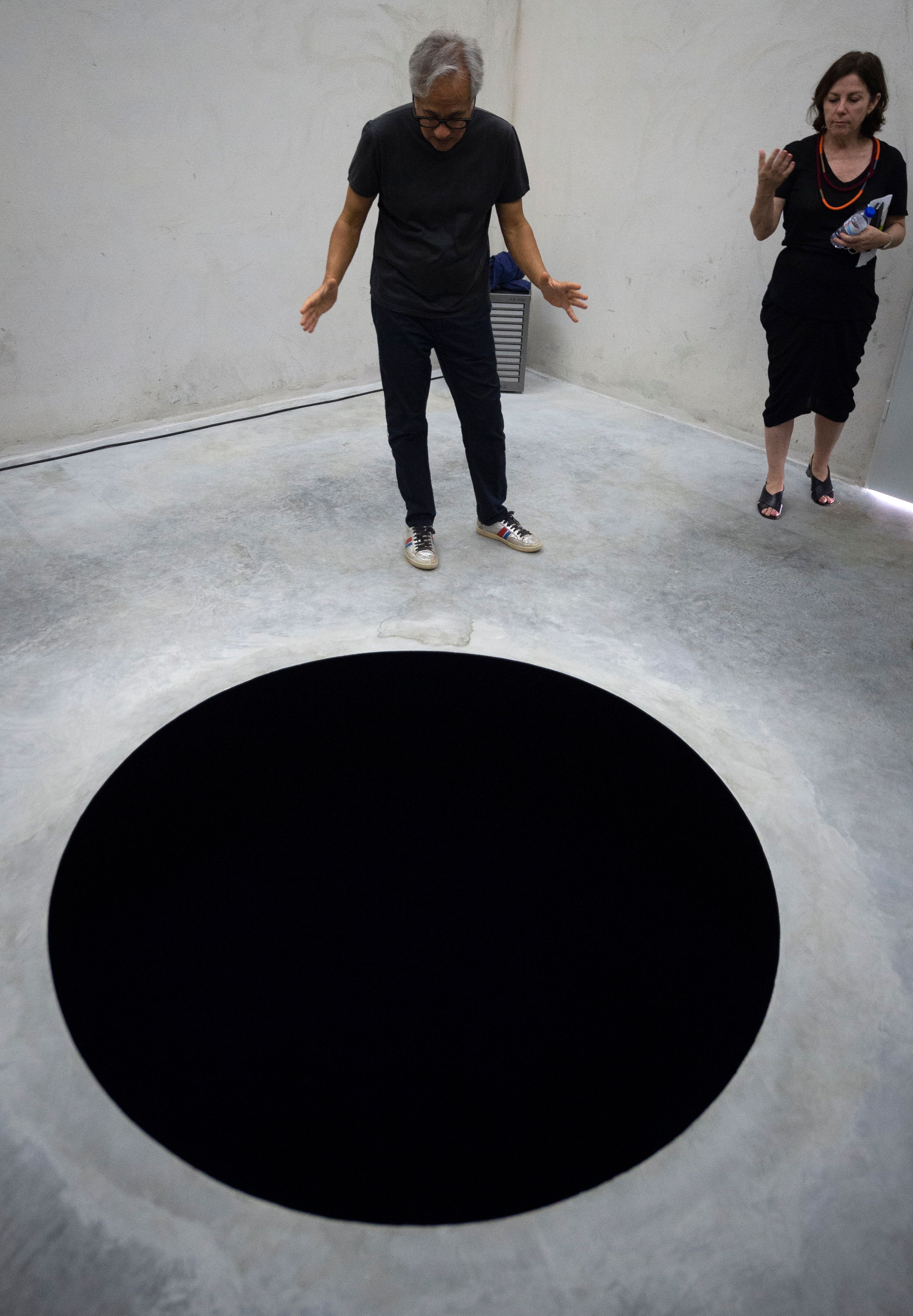 Artist Anish Kapoor and curator Suzanne Cotter stand next to Kapoor's artwork "Descent into Limbo," a circular black void on the floor.