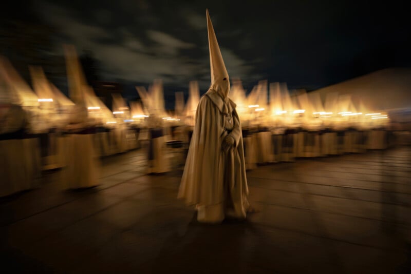 A group of people in white robes and tall, pointed hoods participate in a nighttime procession, holding candles or torches. The image is blurred, giving a sense of movement and solemn atmosphere.