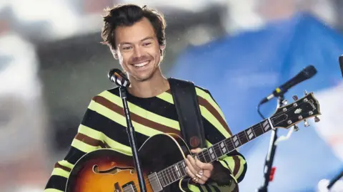GC Images via Getty Images Harry Styles smiles in front of a microphone on a stand onstage as he holds a Gibson Les Paul guitar while wearing a striped green, black and brown top, with background bokeh behind, during a gig in New York in 2022.