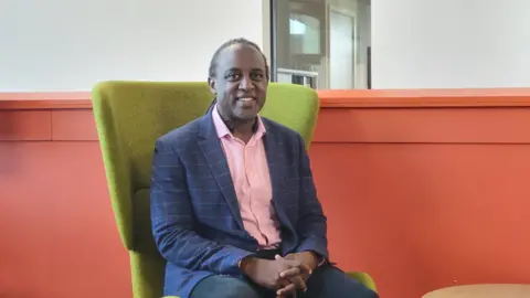 Ulster University Shows a man in a pink shirt, blue and pink jacket and dark trousers sitting in a green chair in front of an orange wall.
