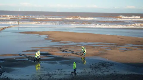 BBC/Joe Bilton People wearing high vis weather gear are walking across the beach.  The tide is out.