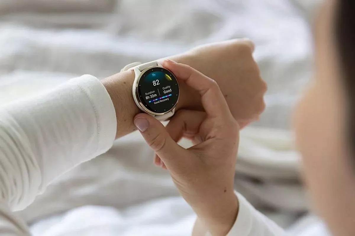 An individual in a white long-sleeved shirt is examining a modern wristwatch with a digital display, held in their hands against a backdrop of neatly arranged white bed linens.