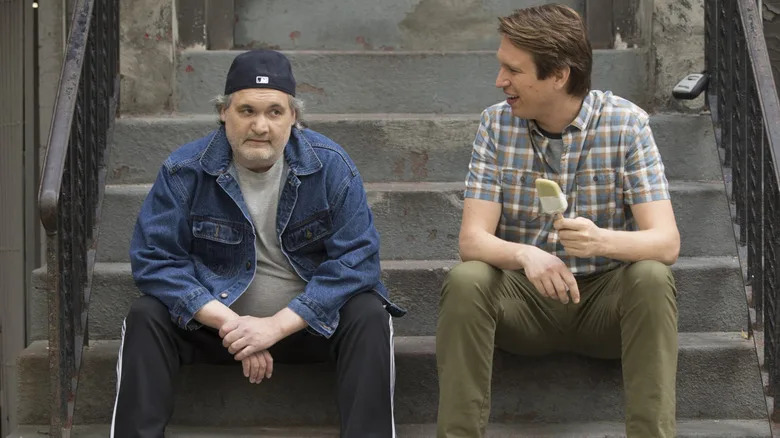 Artie Lange wears a backwards baseball cap while Pete Holmes eats an ice cream while sitting on a stoop in "Crashing"