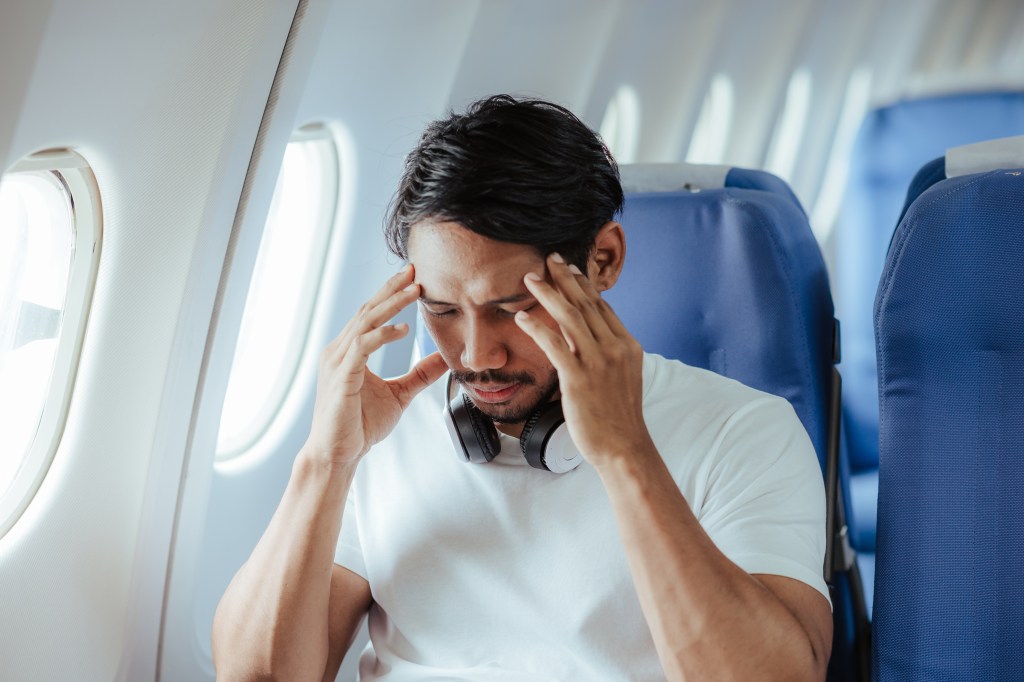 A man in an airplane seat, wearing a white t-shirt and headphones around his neck, holds his temples with both hands, looking down with a pained expression.