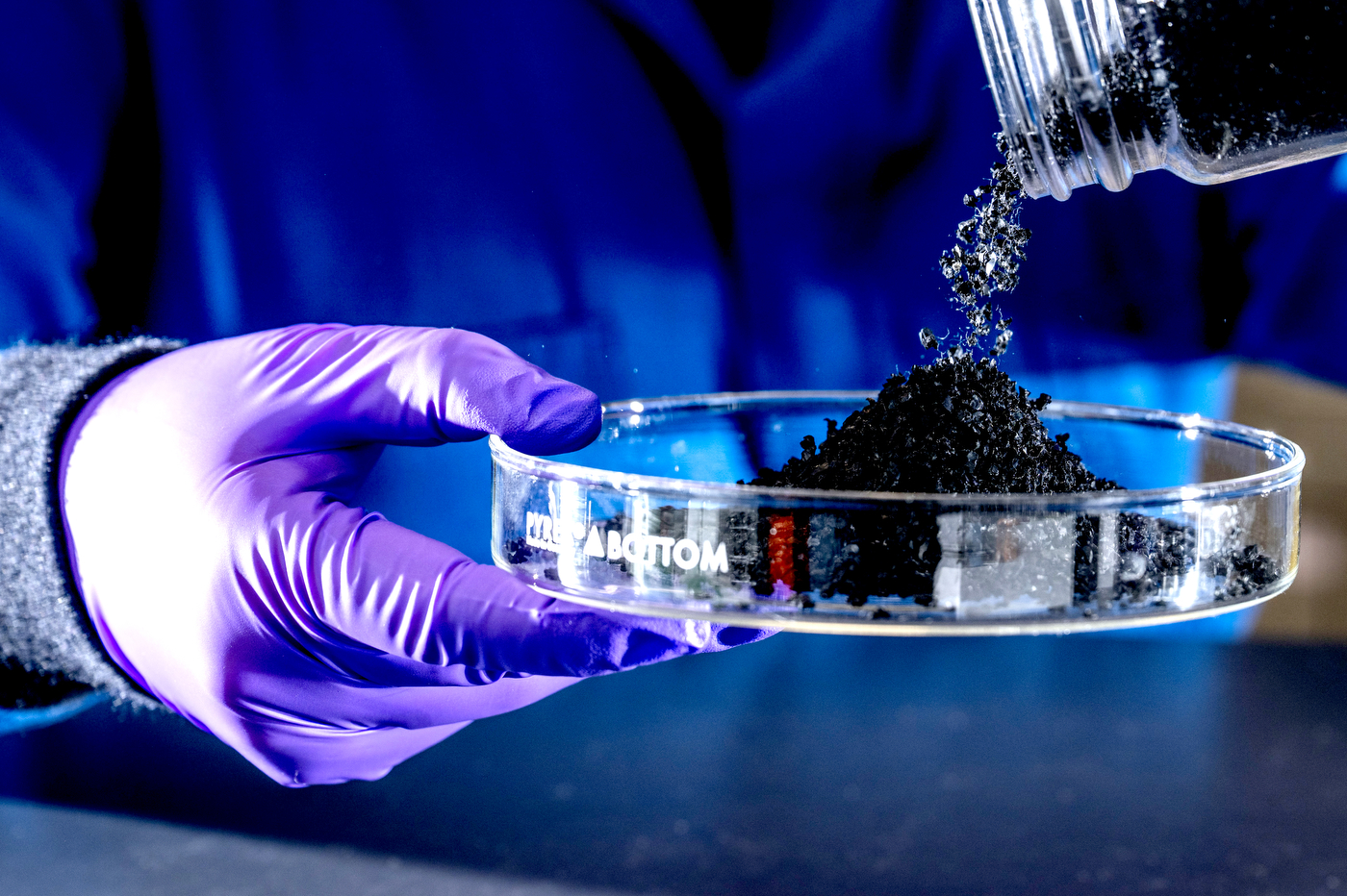 A hand in a latex glove holds a glass dish into which is poured particles of recycled tire rubber.