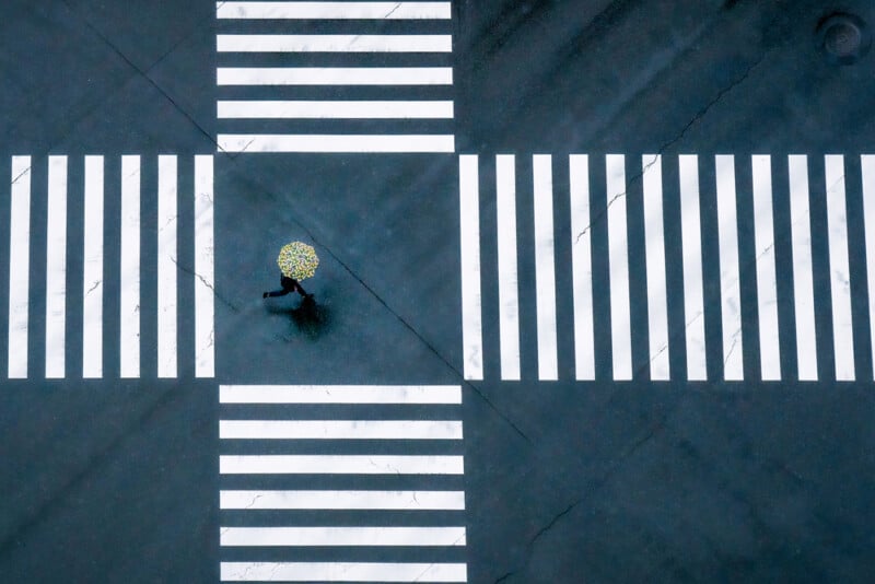 A person holding a yellow umbrella with a floral pattern crosses an empty intersection with wide, white crosswalk lines, viewed from above on a rainy day.