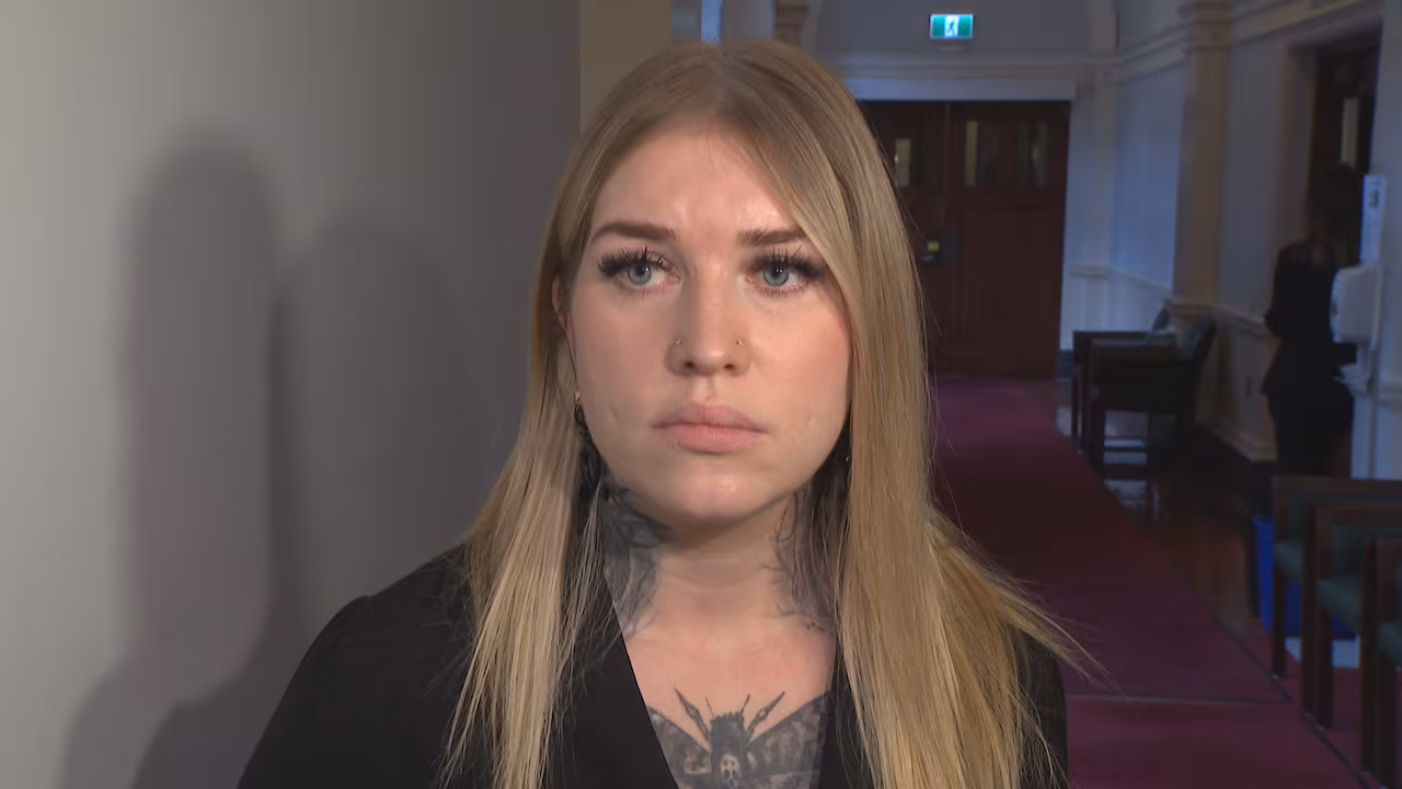 An woman with blond hair and tattoos on her neck is seen in a carpeted hallway.