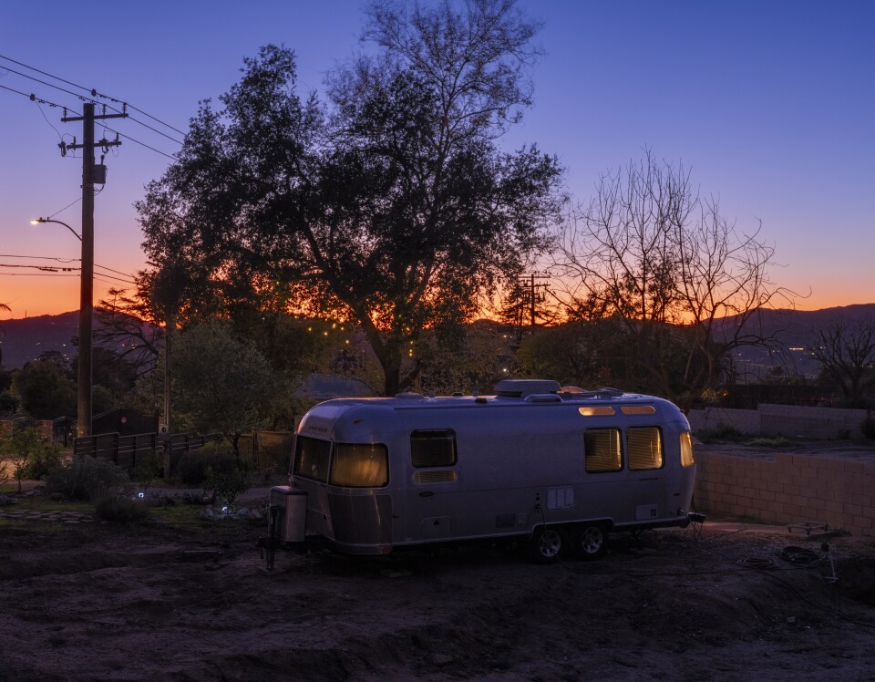A serene twilight scene featuring a classic silver Airstream trailer parked on a dirt lot, framed by silhouetted trees and a vivid sunset sky.