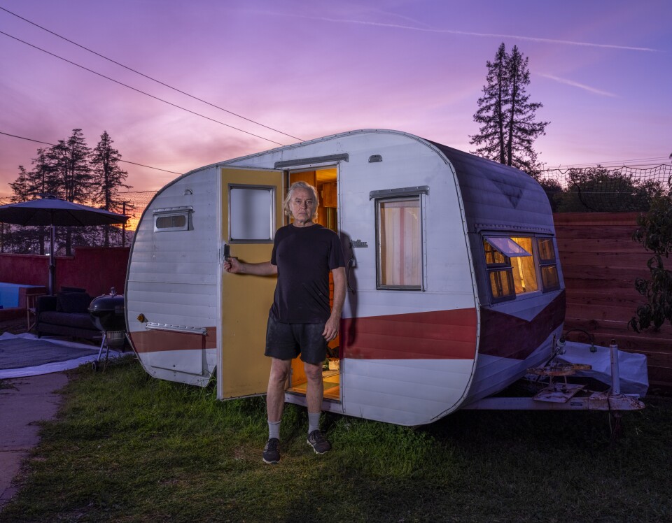 A vintage-style travel trailer parked on a grassy area during twilight, with a vivid purple and pink sky in the background. A man in shorts and a t-shirt stands at the door.
