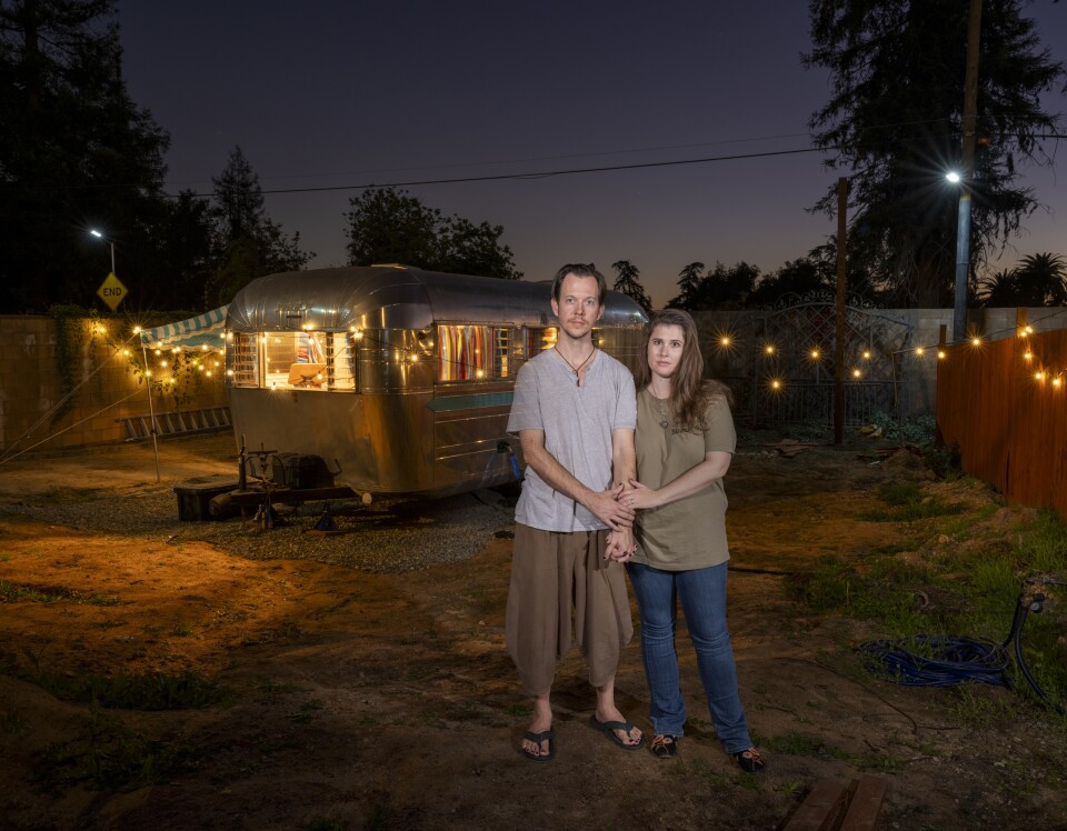 The image shows an outdoor nighttime scene featuring a polished silver Airstream trailer illuminated warmly from within. The trailer is parked on a dirt or gravel lot, surrounded by string lights that create a cozy, festive atmosphere. In the foreground, two people stand close together, holding hands, positioned slightly off-center in front of the trailer.  