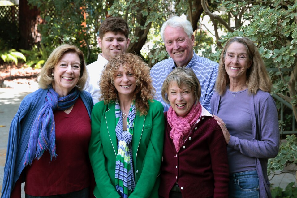 A staff picture of the "Palisadian-Post". Six people stand together, four people are in front and two are in back. The person at the far left and foreground wears a light blue jacket and scarf and a red shirt. The person in the middle foreground wears a green blazer and a green and blue scarf. Another person stands to her side and wears a pink scarf and dark red shirt. On the right side of the picture a woman wears a purple shirt, jeans and a purple cardigan. In the background, on the left side, one person wears a white button up shirt. To the side of him Bill Bruns wears a white and blue striped button up shirt. They all smile and pose for a camera. They are in a garden surrounded by green shrubbery.
