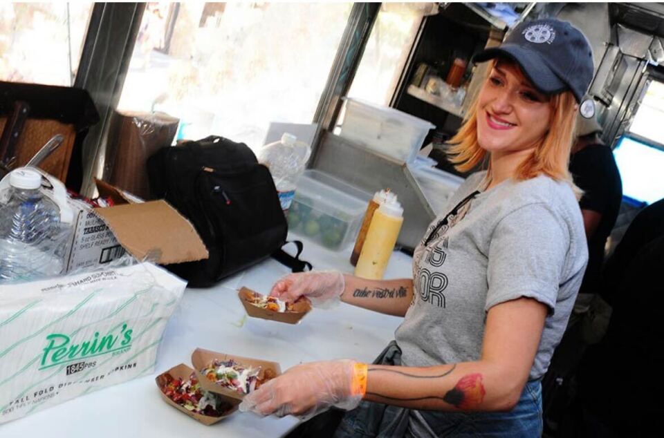 A woman in a dark blue hat wears food gloves and handles small baskets of food inside a food truck. She wears a gray shirt and light blue jeans. Napkins, a black bag, and condiments can be seen on the table in front of her. Behind her a person in a black shirt is working.