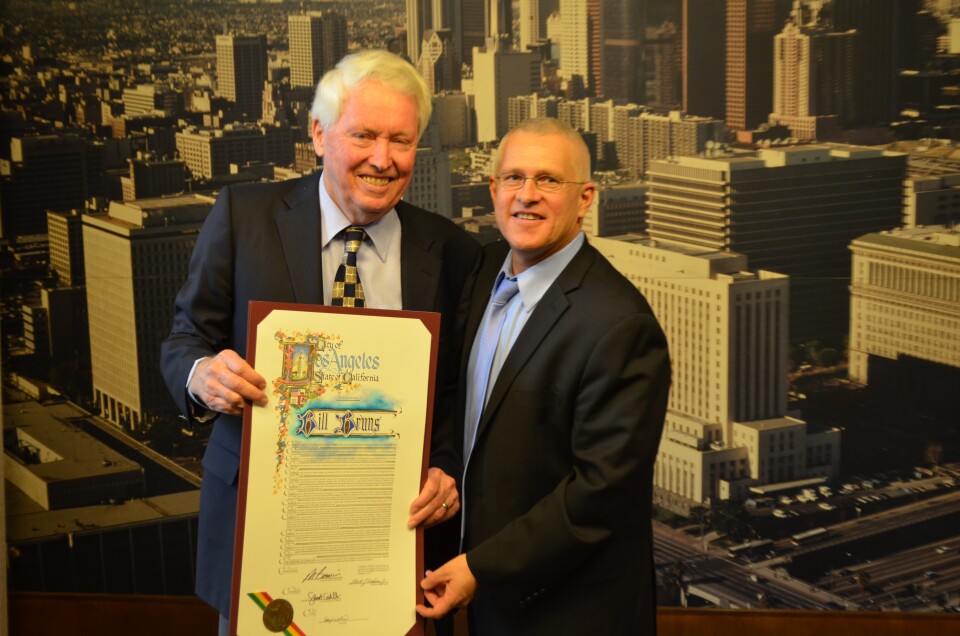 A man in a blue suit wearing a blue collared shirt and a blue and tan tie stands next to another man with glasses, a black suit, a blue collared shirt and a light blue tie. An aerial view photograph of downtown Los Angeles can be seen behind them. The man in the blue suit holds an honorary certificate up.