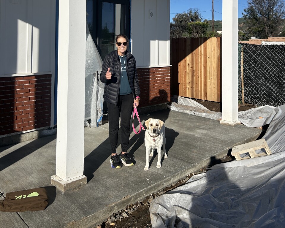 A woman in a blue puffer jacket, a sweatshirt, jogging pants and running shoes stands in the construction site of a home with a white dog on a leash. She wears sunglasses and makes a thumbs up gesture to the camera.