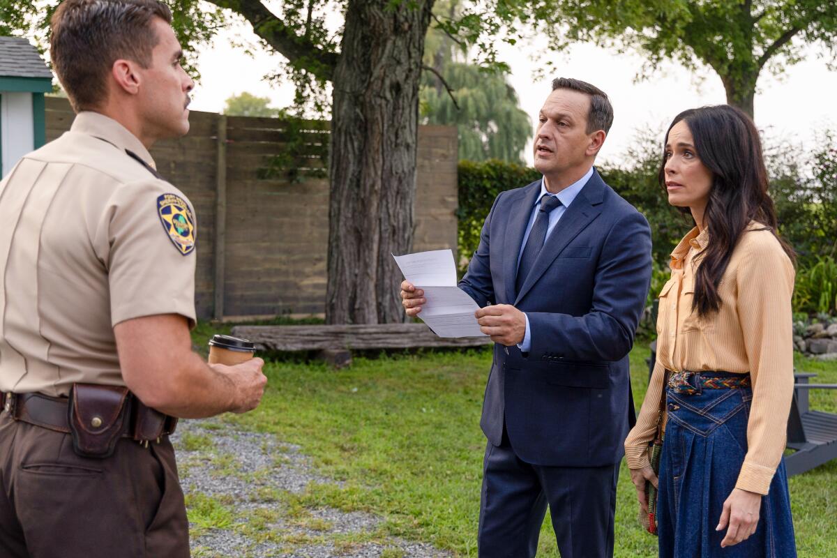 A sheriff in a tan uniform looks at a man in a blue suit and a woman an orange top and denim skirt standing next to him.
