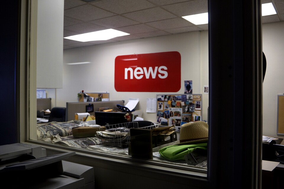 A room is seen through a window. A red sign with white letters that says "news" sits in the middle of the room. Old newspapers can be seen on the other side of the windowpane. Next to the newspapers are a basket and a straw hat. A copy machine is seen in front of the window. Desks and a bulletin board filled with pictues can be seen beyond the window in the room.