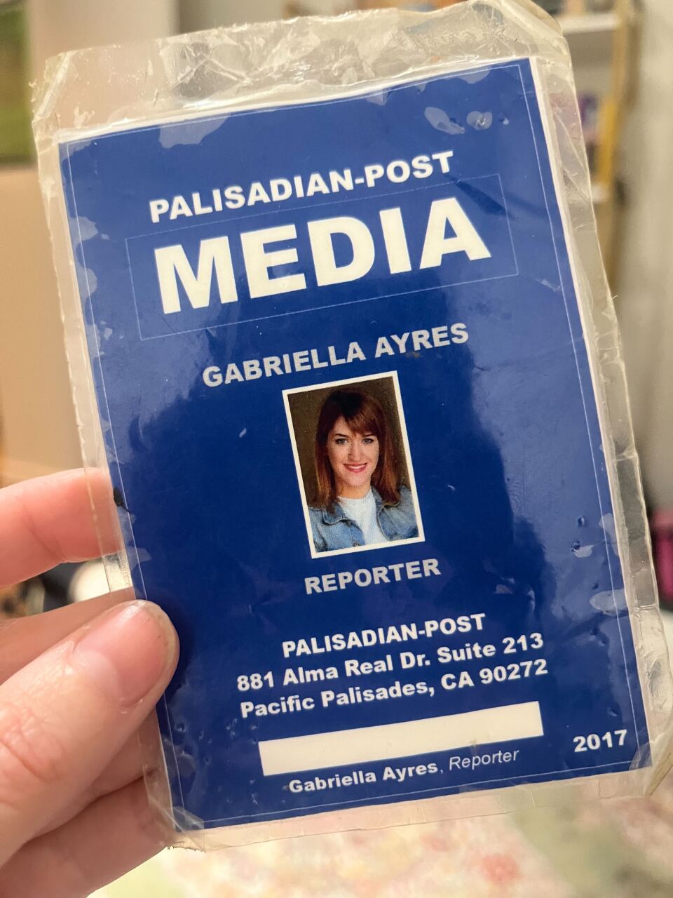 A dark blue laminated "media pass" is seen. The media pass depicts a woman with brown hair in a white T-shirt and blue sweater. Underneath the picture is written the word "reporter". Above the picture the words "Gabriella Ayres" is written. Above those words "Palisadian-Post Media" is written.