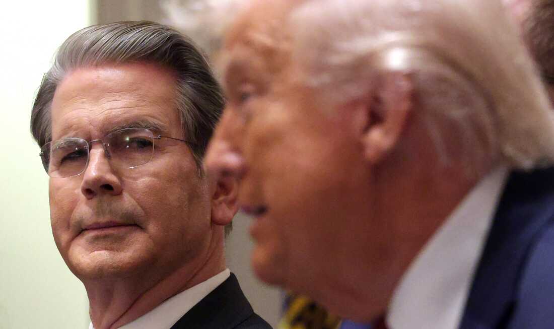 Photographed from the neck up, Treasury Secretary Scott Bessent is on the left side of the frame, looking in the direction of President Trump, who appears in profile on the right side of the frame.