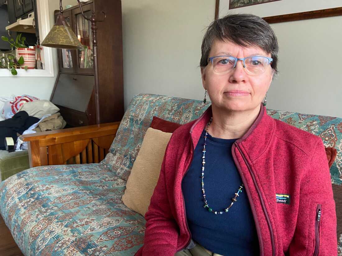 Mary Cowhey sits on a sofa. She's wearing a blue shirt, a cranberry-colored jacket, a necklace and glasses.