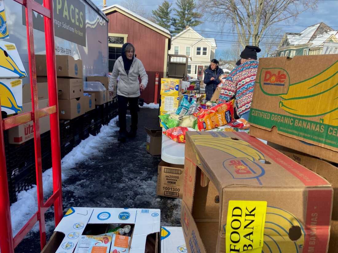 Volunteer Ramona Kallem, wearing a hoodie, walks near tables set up outdoors as part of a pop-up food bank. On the tables are groceries such as bags of rice cakes and canned foods. Cardboard boxes are piled near the tables, and slushy snow covers some of the ground.