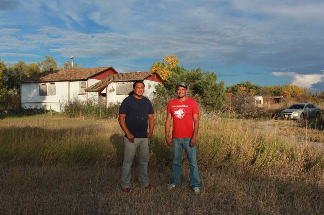 Lonny and Teyon Fritzler stand outside their childhood home. Plywood covers the windows of the white house, and tall gold-colored grass is high in the yard between the brothers and the house.