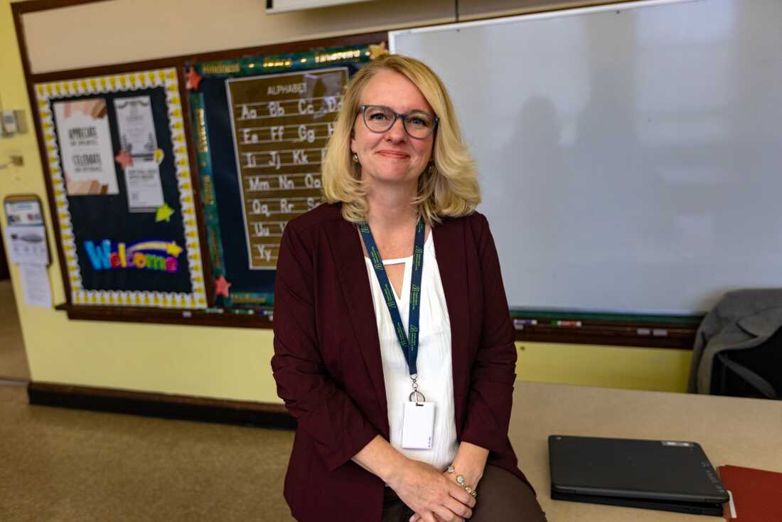 A woman sits at the edge of a table with one hand atop the other in her lap. She's wearing glasses and has an ID lanyard around her neck.