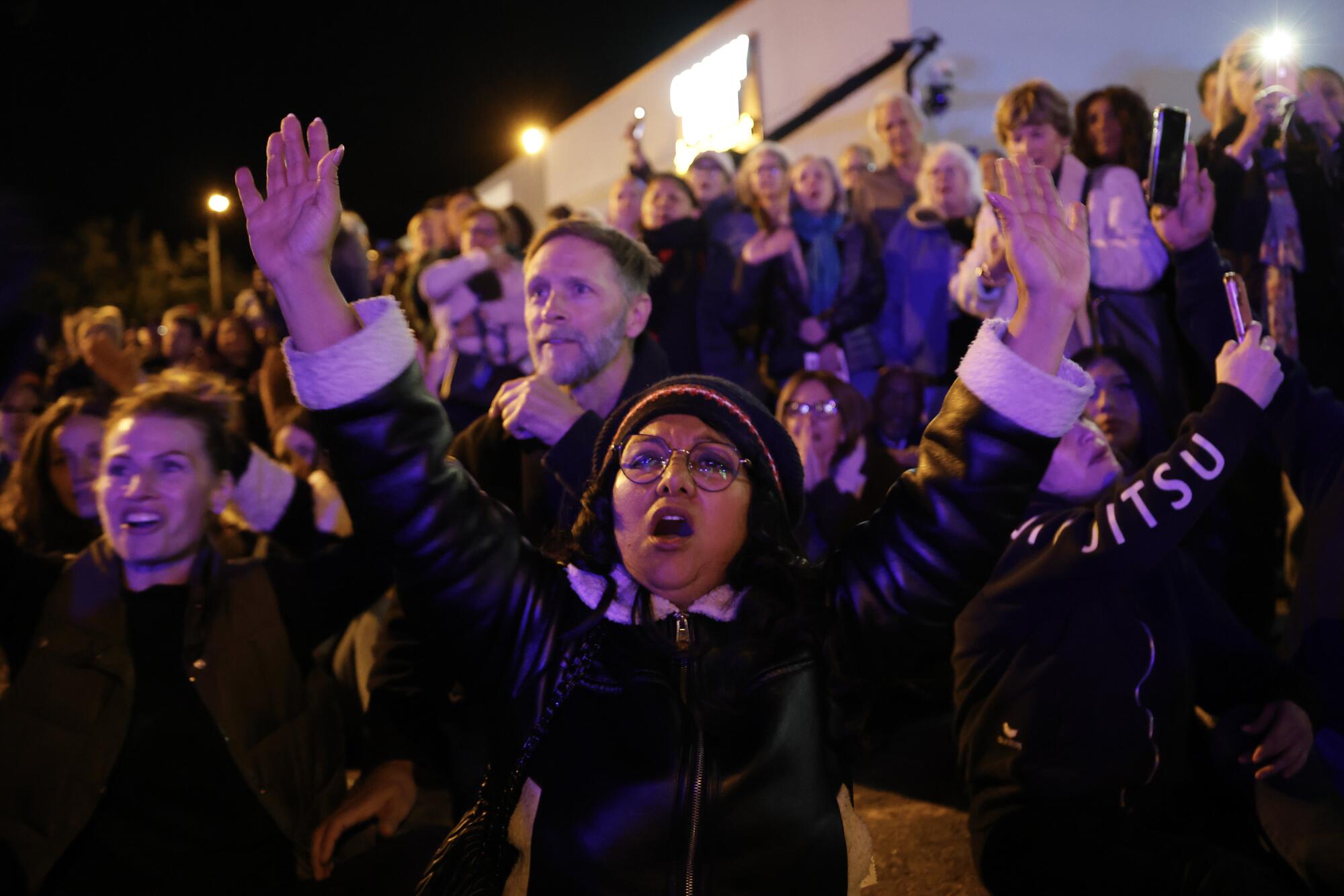 A woman among a crowd holds up both arms.